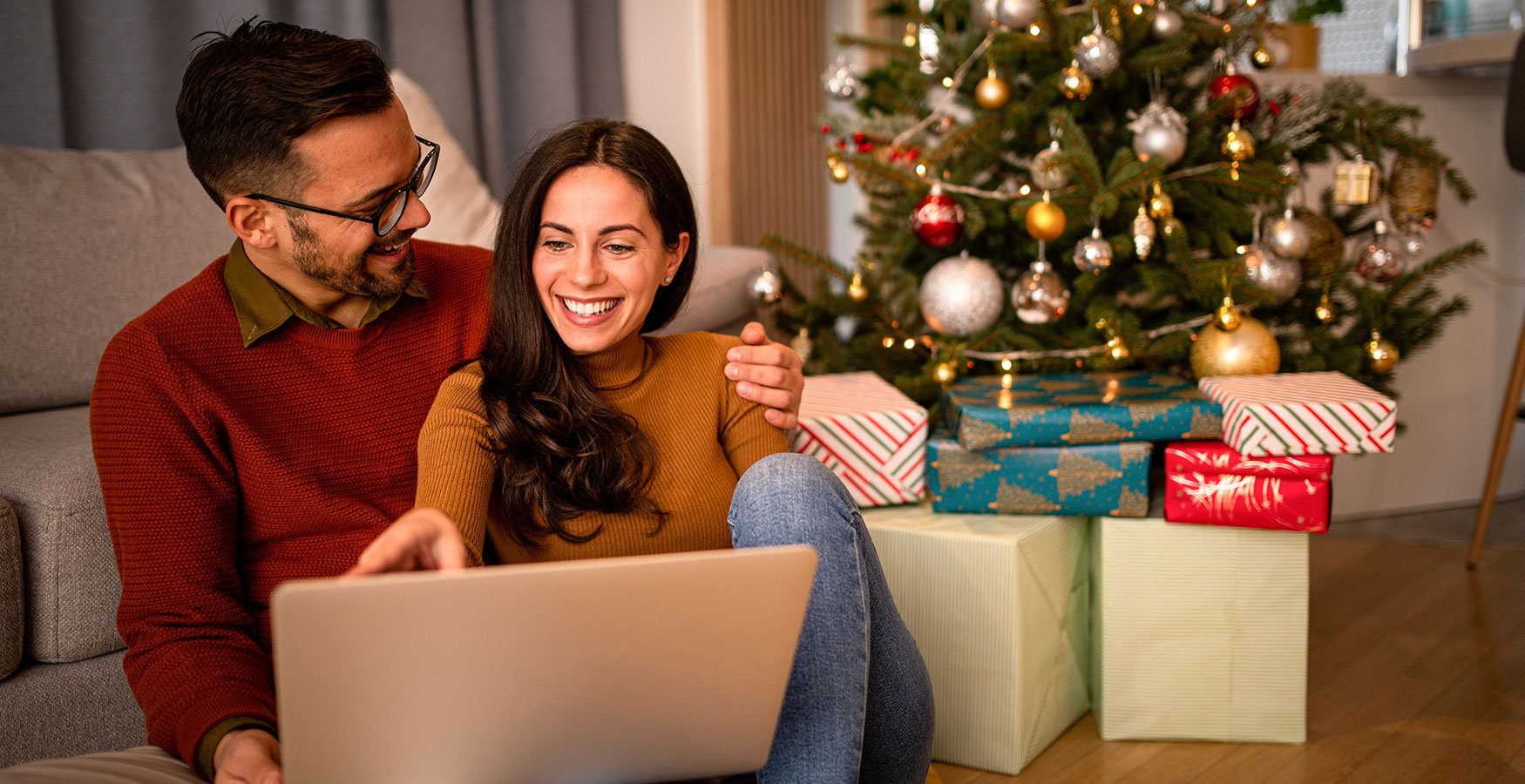 Happy couple sitting in front of Christmas tree with presents under. Looking at their laptop. 