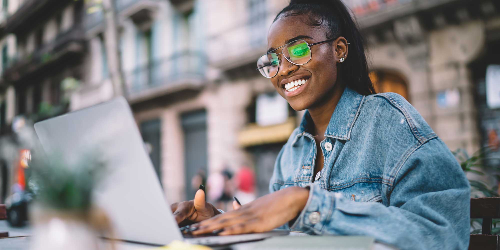 College girl wearing a denim jacket, smiling at her laptop.