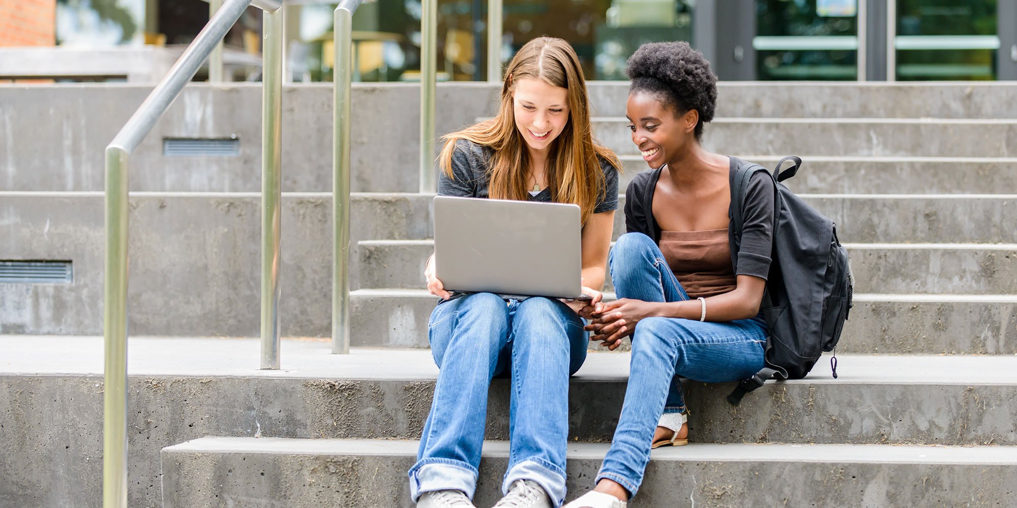 Two girls sitting on stair steps looking at a laptop. 