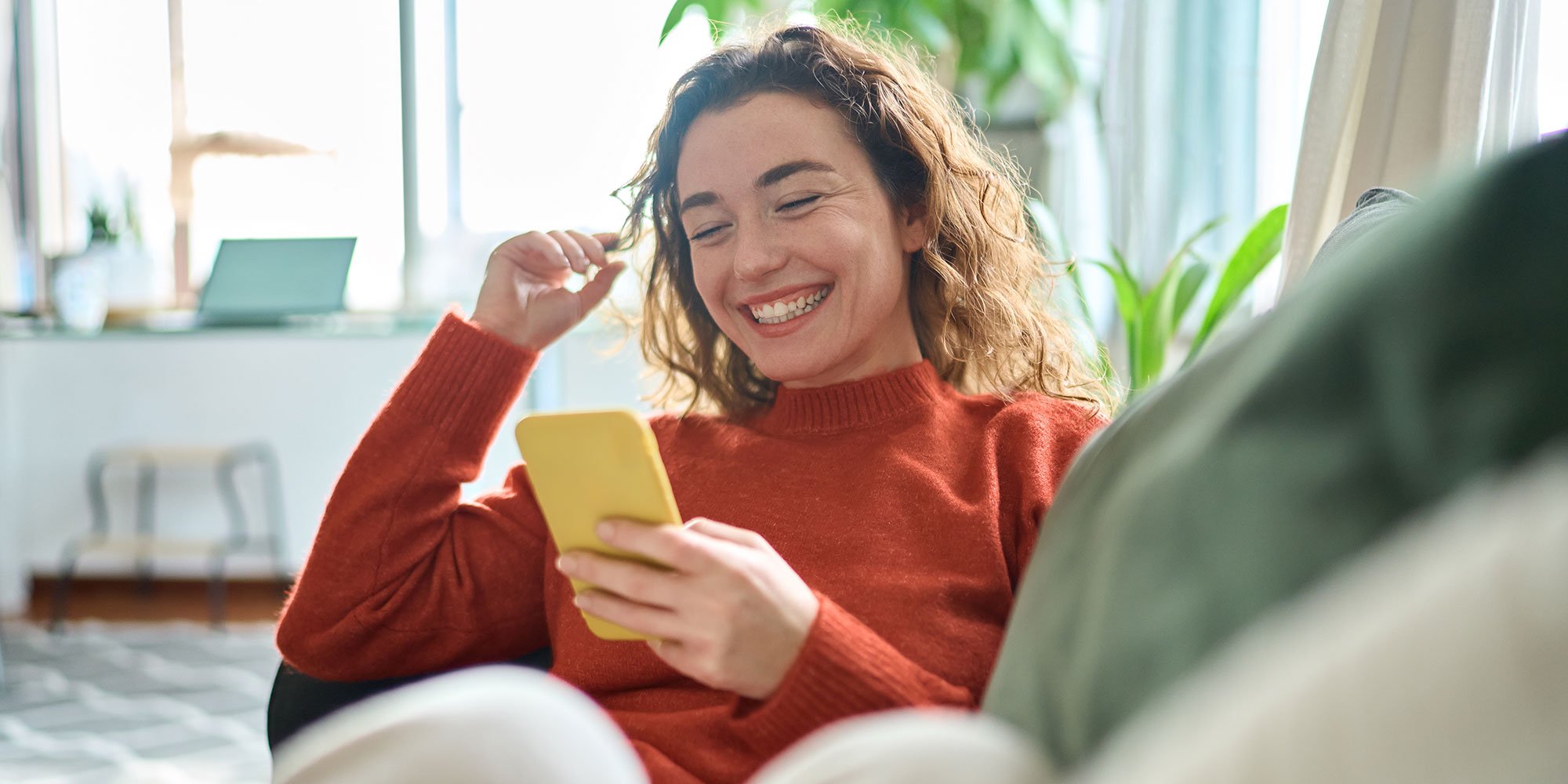 Woman smiling while looking at a smartphone on a couch.