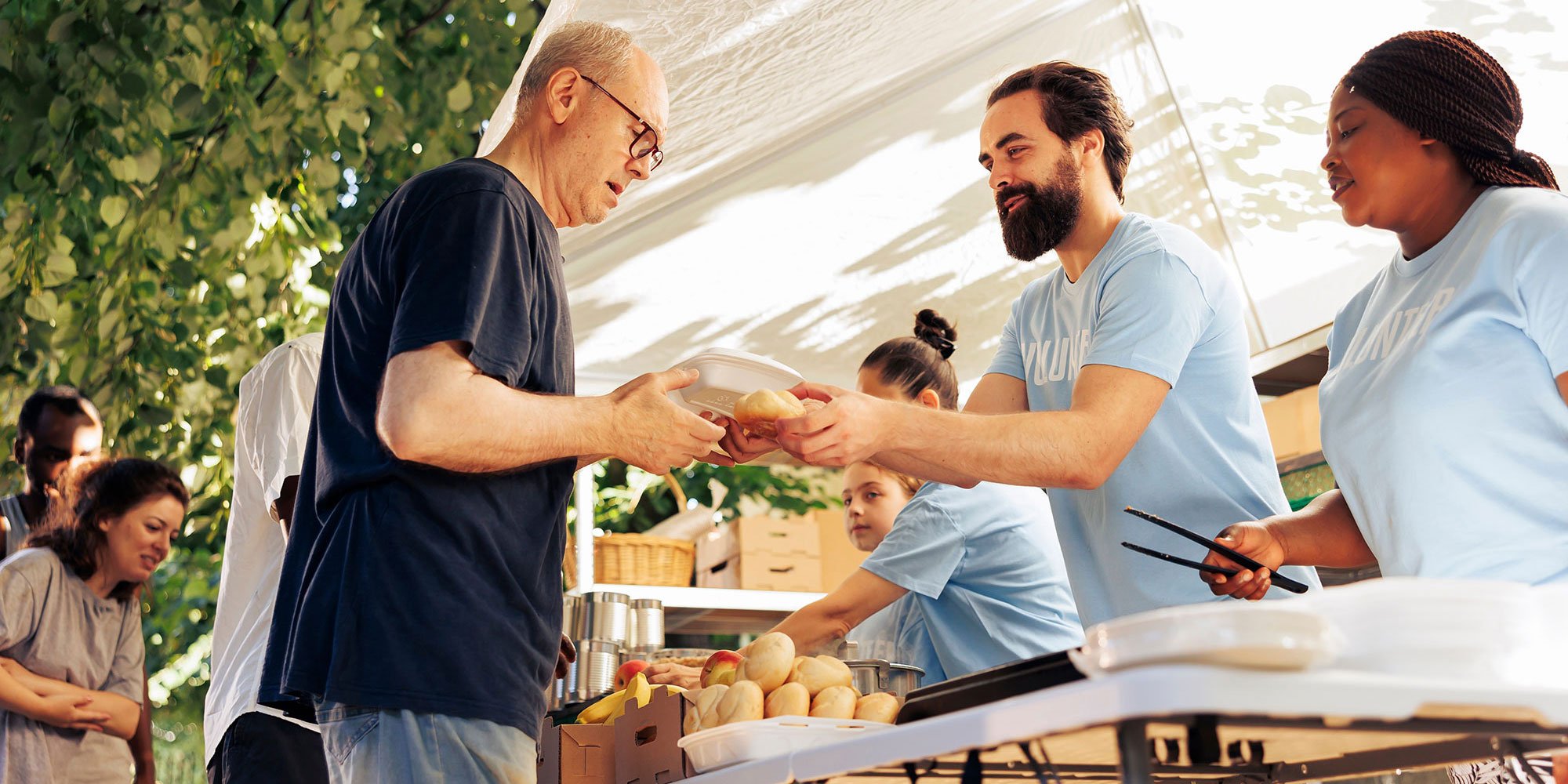 Volunteers in blue shirts handing a meal to a man at an outdoor food distribution event.