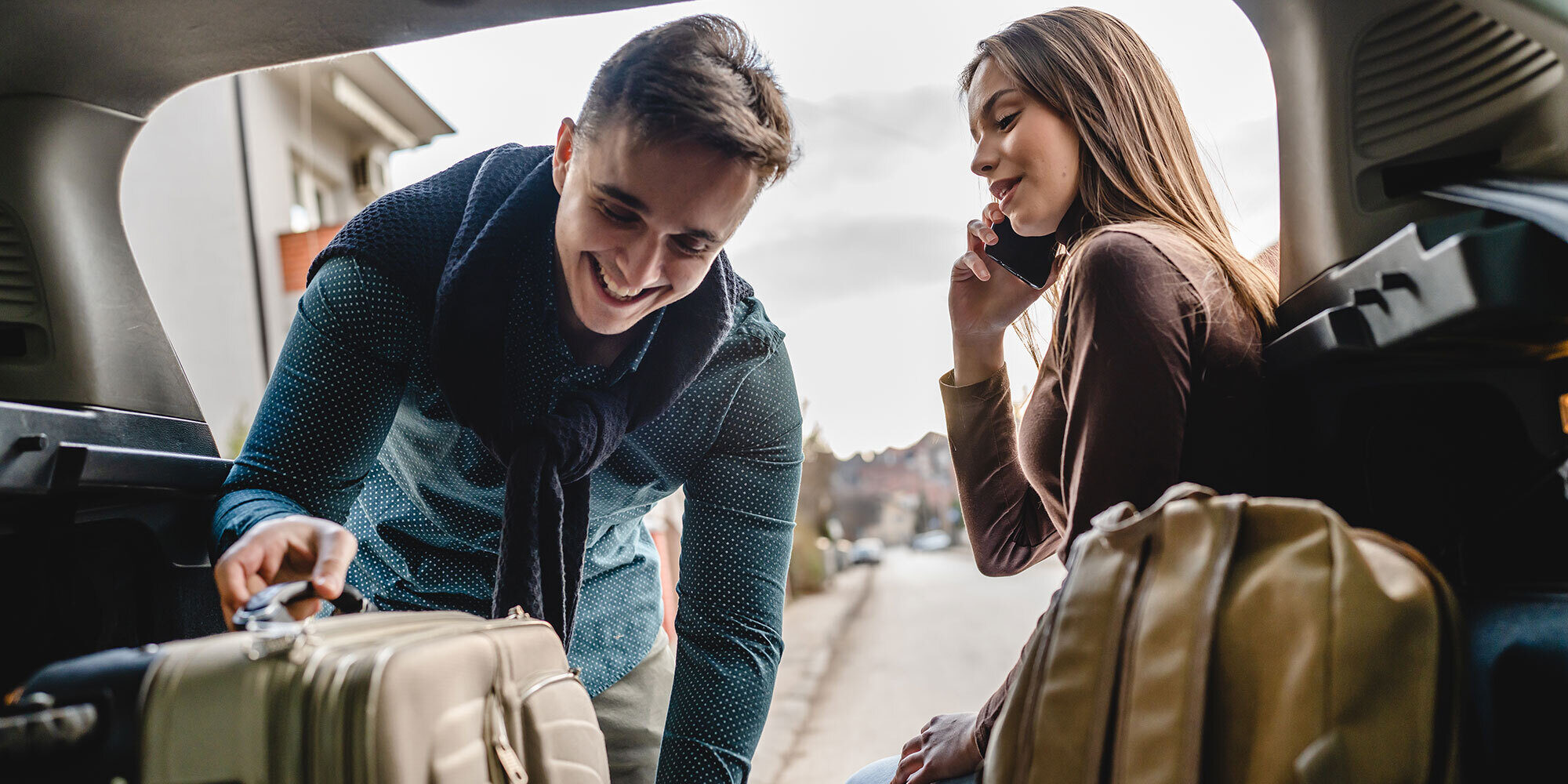 Couple outside their car. Woman is on the phone, man is grabbing luggage.