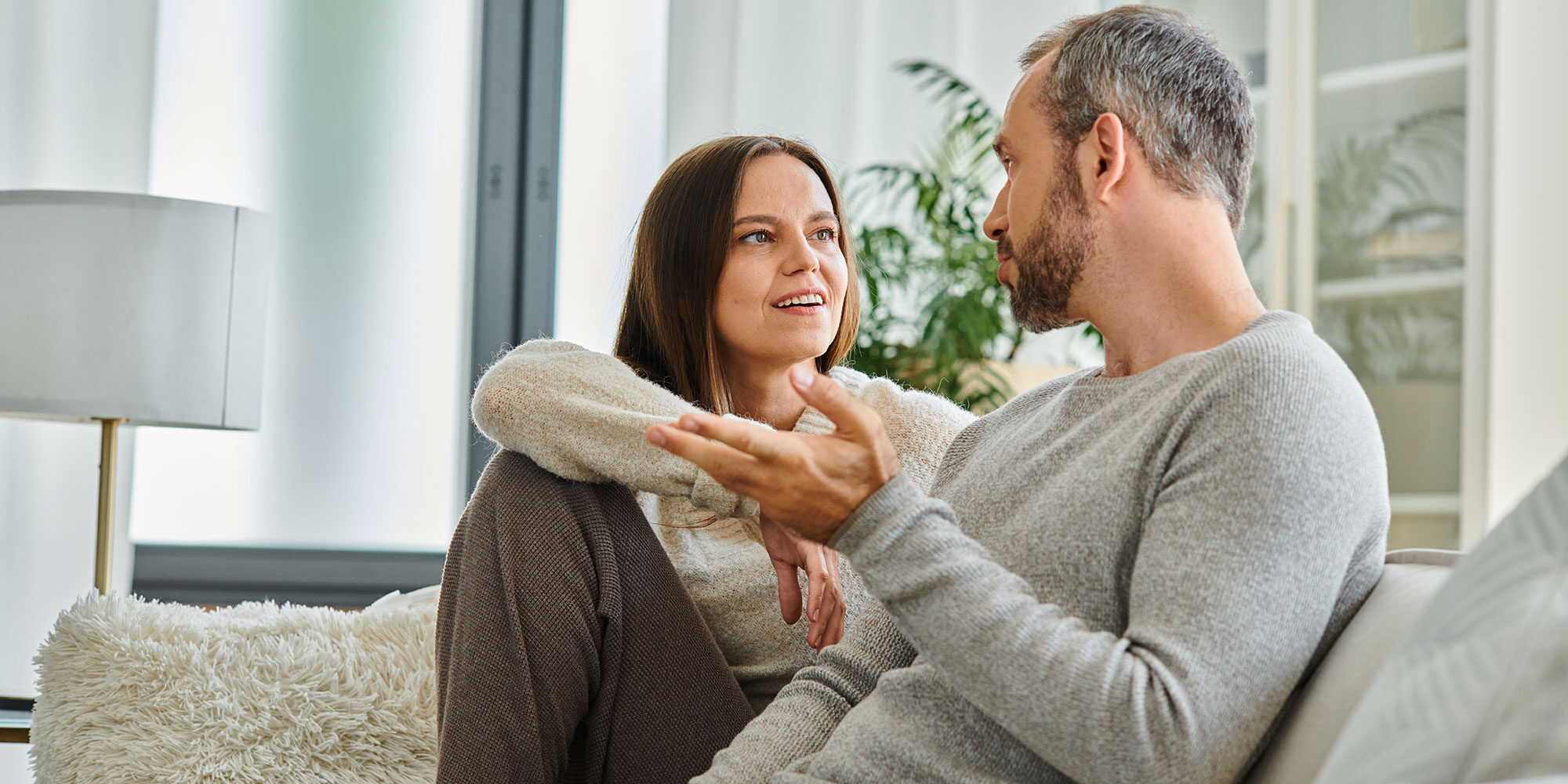 A couple sits on a couch, engaged in a conversation, with the woman smiling and listening attentively while the man gestures with his hand.