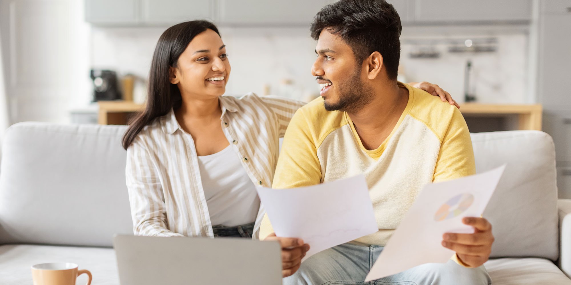 A smiling couple sits on a couch with a laptop and financial documents, discussing their finances in a bright kitchen setting.