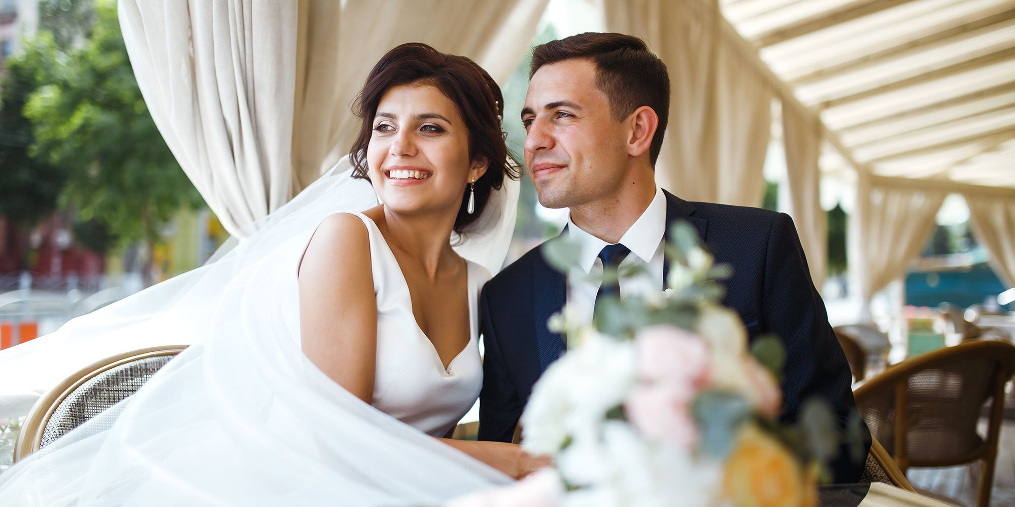 A smiling bride and groom sit together under a decorated canopy, looking happy and excited on their wedding day.