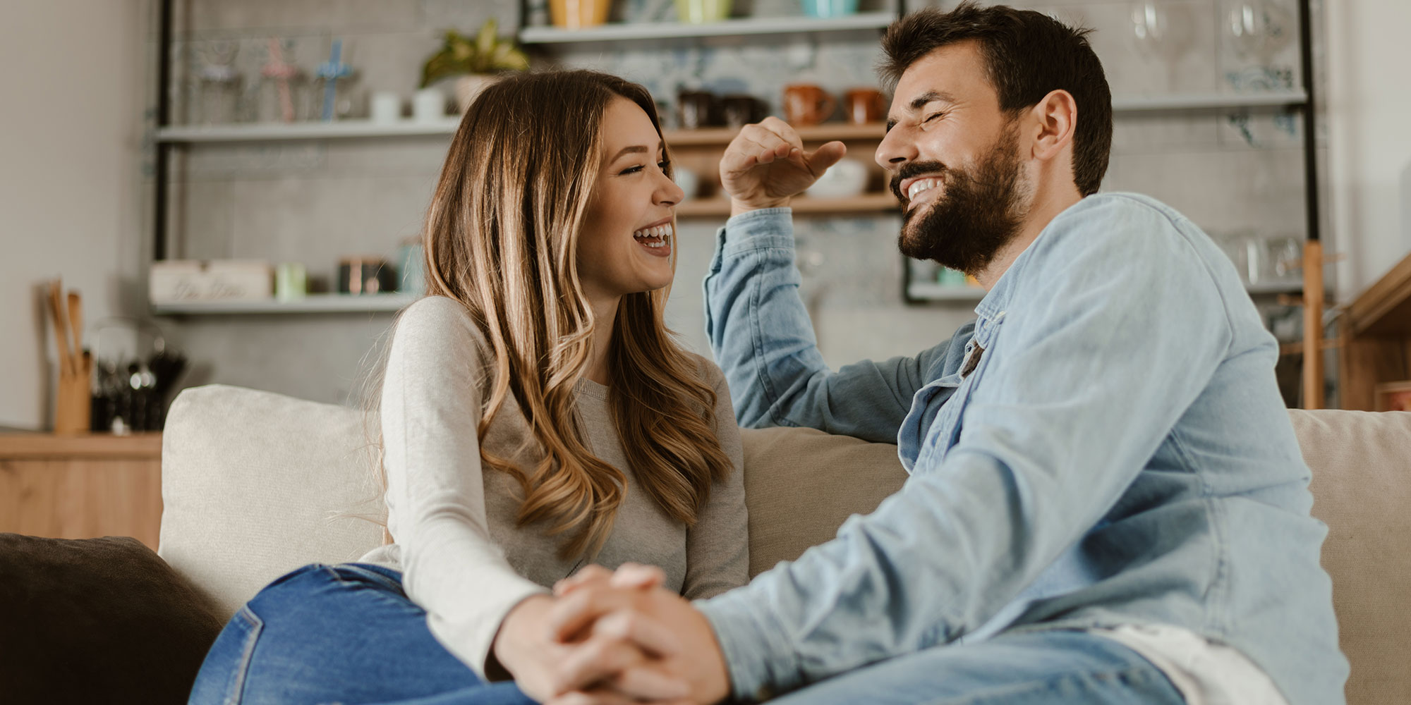 A smiling couple sits on a couch, holding hands and laughing together in a cozy kitchen setting.