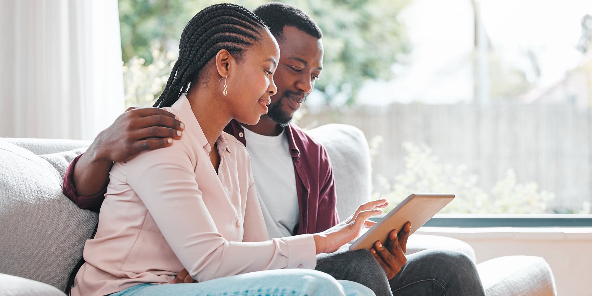 A smiling couple sits on a couch, looking at a tablet together, with the man’s arm around the woman.