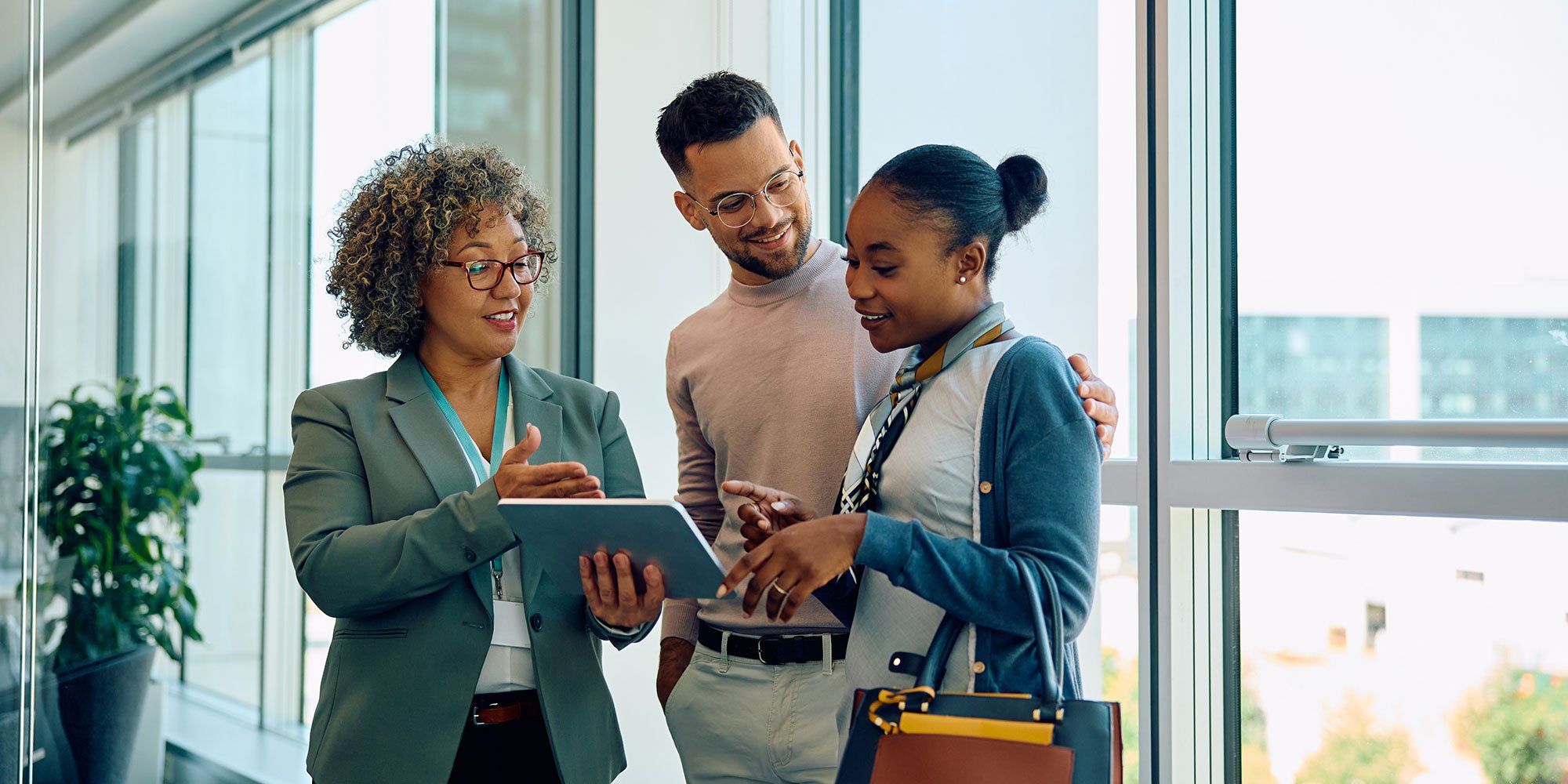 A professional woman in a blazer holds a tablet and discusses financial details with a smiling couple in a bright office setting.