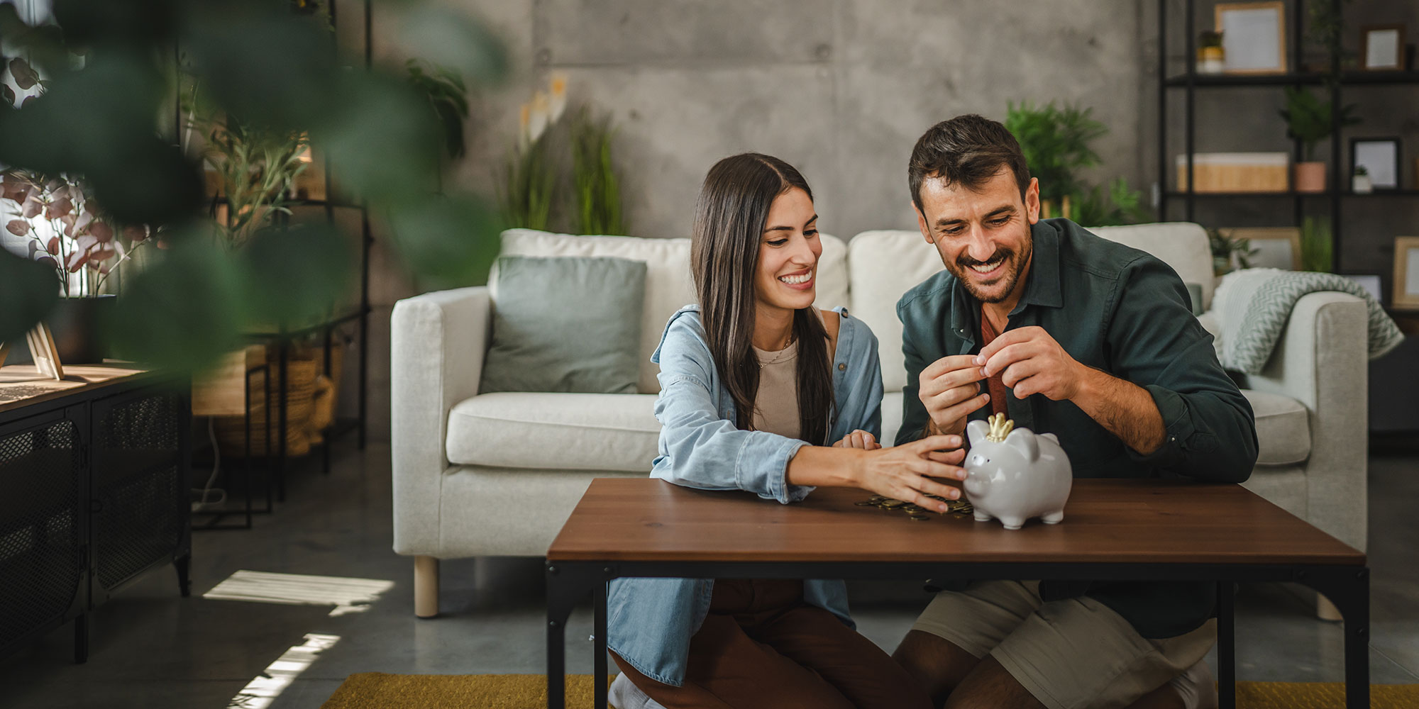 Smiling couple sits on the floor, saving money in a piggy bank in a cozy, modern living room.
